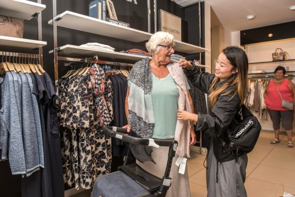 Vrouw helpt een oudere vrouw met het uitkiezen van kleding in een winkel.