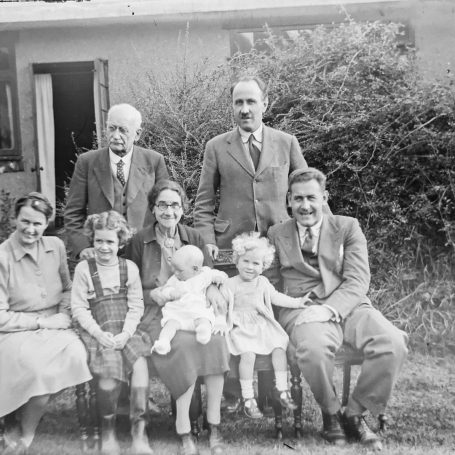 Zes mensen en een kind met een pop poseren voor een huisje in de tuin.