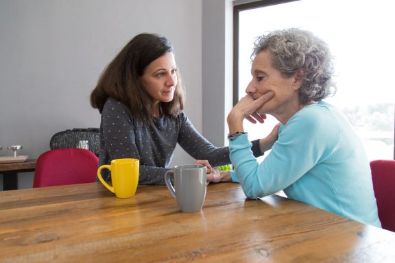 Twee vrouwen zitten aan een tafel en hebben een gesprek, met koffiekopjes voor hen.