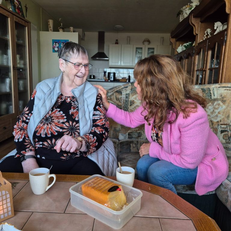 Een lachende vrouw en een oudere dame zitten samen aan een tafel met koffie en gebak.