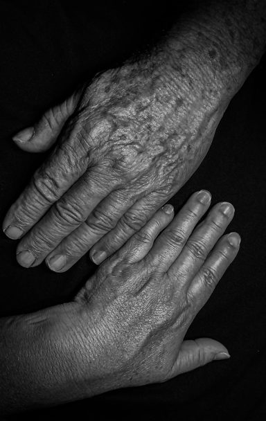 Twee handen, een oudere met zichtbare rimpels en een jongere, op een donkere achtergrond.